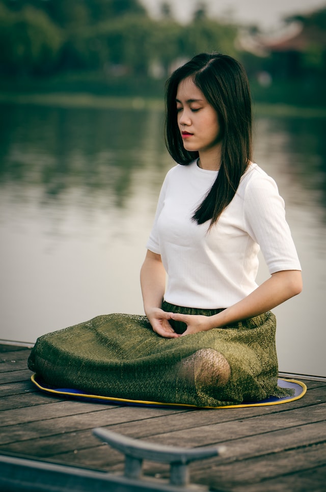 young adult female sitting in meditation pose on a dock with lake blurred out in background.