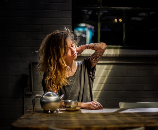 Young woman with tattoos on her arm sitting at a table with tea cup and pot. She is rubbing her stiff neck.
