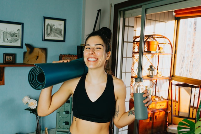 Young women with yoga mat rolled up on her shoulder, water bottle in had, happy with very big smile. Standing in her home with flowers in vase and framed pen drawings on wall